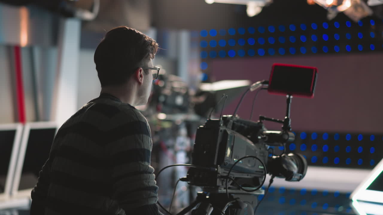 Camera operator adjusting professional broadcast camera on tripod in high tech studio environment with blue LED lights background blurred monitors cables visible focusing on filming process