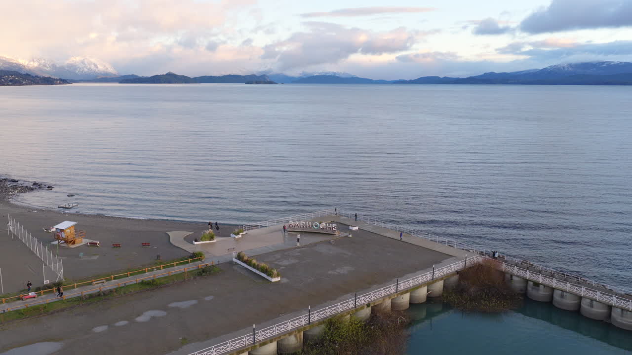 Slow drone glide above pier extending into Nahuel Huapi Lake with distant snowy mountains