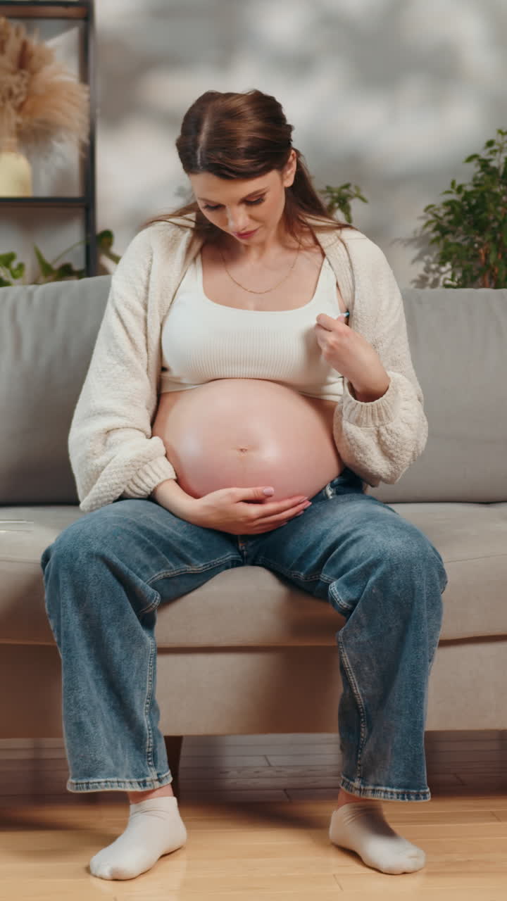 Expecting mother cradles large belly while sitting casually on sofa looking down with gentle