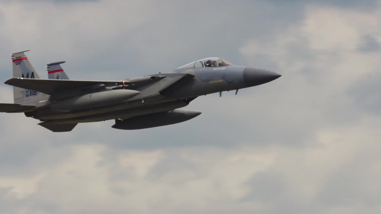 An F-15 fighter jet flies low over forested hills and into an overcast sky.