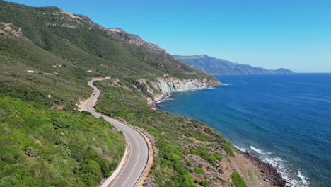 Camino-Costero-Entre-Montañas-Verdes-Y-Mar-Azul-En-Cerdeña,-Italia---Pedestal-Aéreo-4k