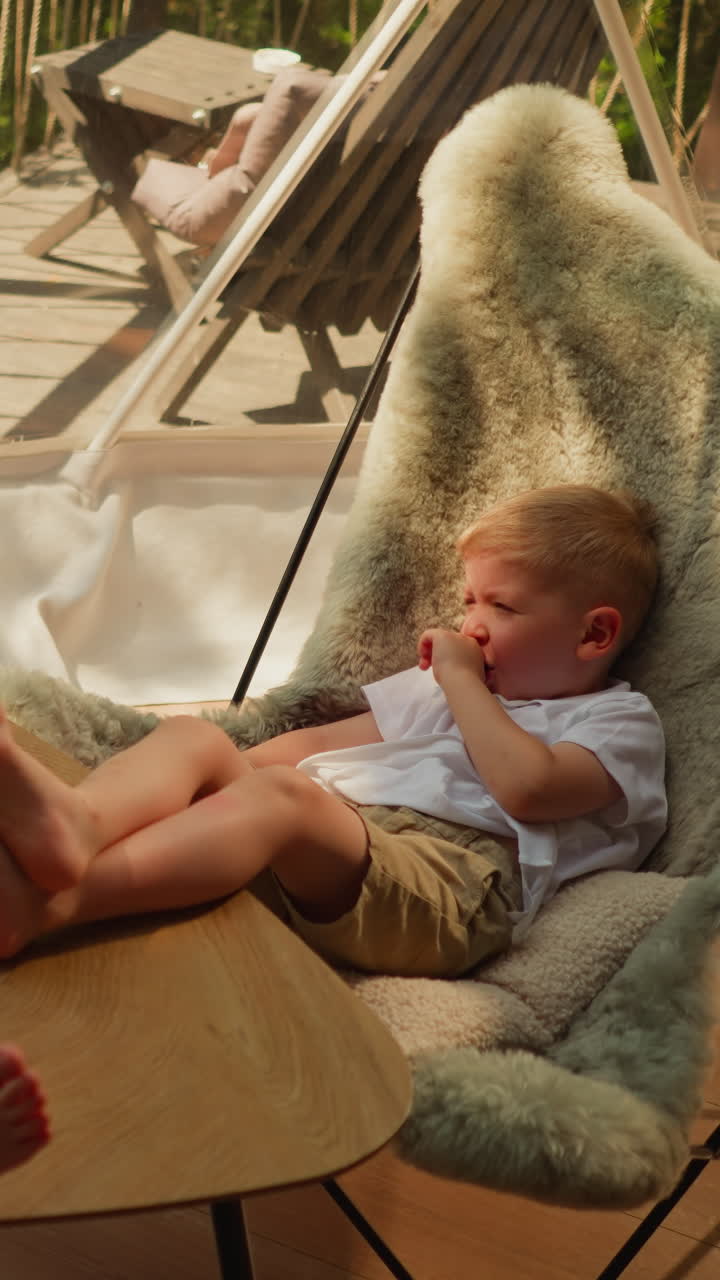 Little children relax on shady glamping ground. Dreaming little boy with girl sit on chairs with fur coatings under glass sphere. Kids at resort