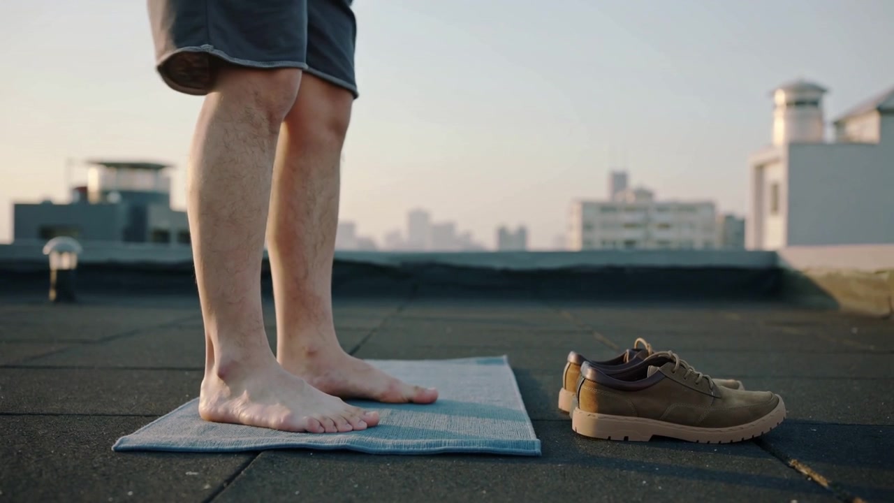 暗い色のショートパンツを履いた男性が、ストライプの屋上マットの上で体を動かしながらバランスを試し、茶色の靴に手を伸ばす。
