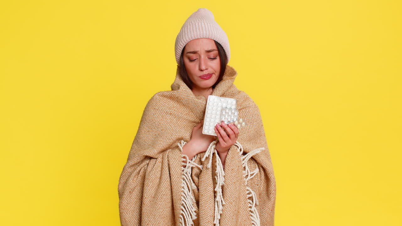 Woman wrapped in blanket wearing hat holding pills thoughtfully looking at them cold flu treatment