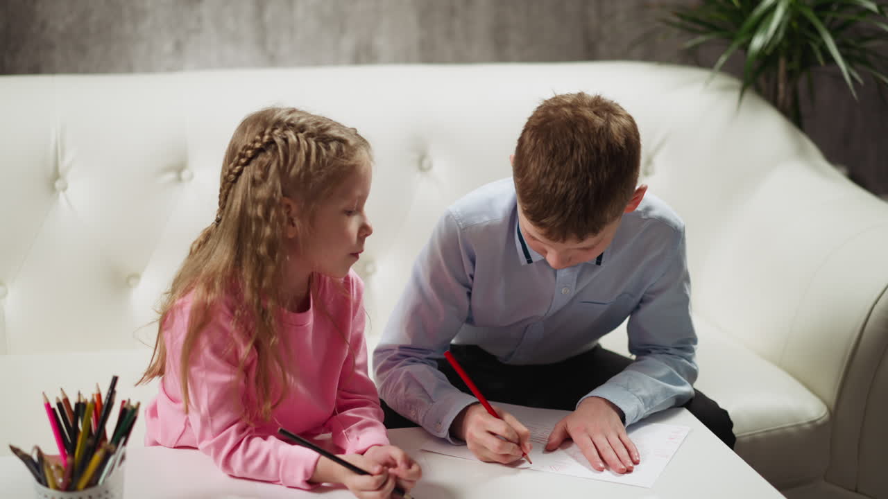 маленькая девочка смотрит на брата, рисующего узоры на бумаге.