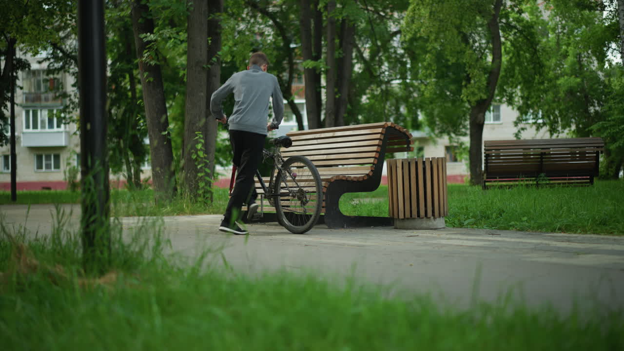 Young boy rides his bicycle through a serene, tree-lined park, he stops by a wooden bench, resting his bike on the seat, the backdrop includes trees, greenery, and a residential building