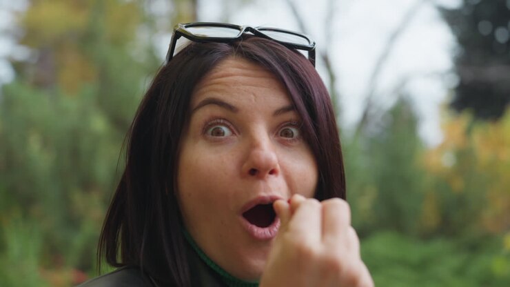 Donna che assaggia una bacca selvatica nella natura, giacca di pelle e occhiali da sole appoggiati, foresta autunnale sullo sfondo con arbusti frondosi, primo piano che mostra sorpresa e poi un sorriso caloroso, momento di vita all'aria aperta spontaneo