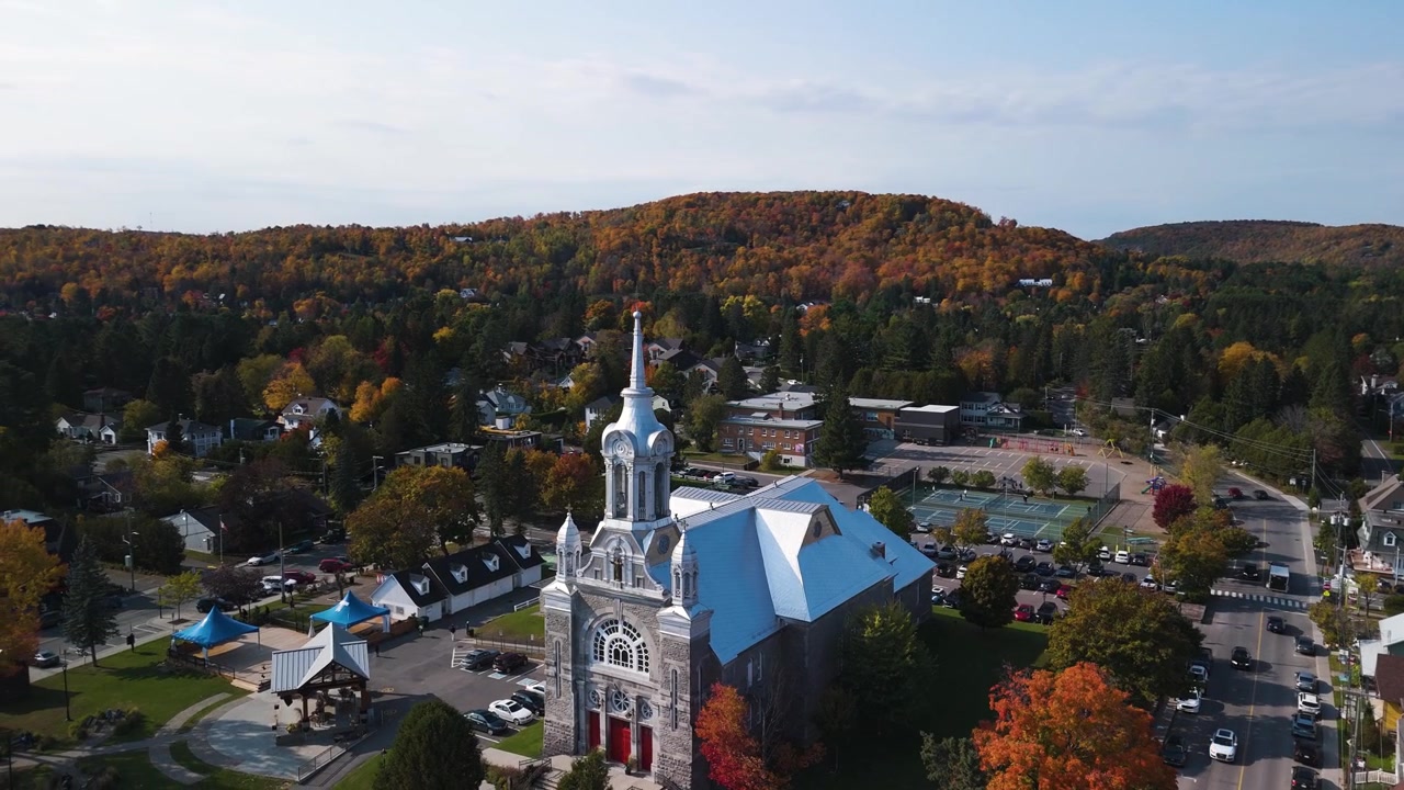 Drone berputar mengelilingi katedral utama di kota Saint-Sauveur, Munisipalitas Regional Les Pays-d'en-Haut, Quebec, Kanada, dengan lanskap pegunungan yang tertutup dedaunan oranye selama musim gugur