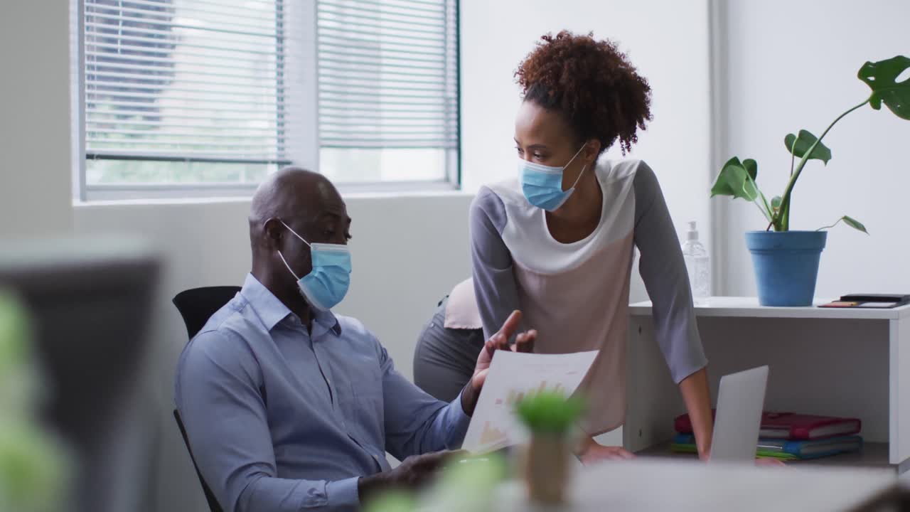 Diverse businessman and businesswoman in face masks discussing and using laptop in office