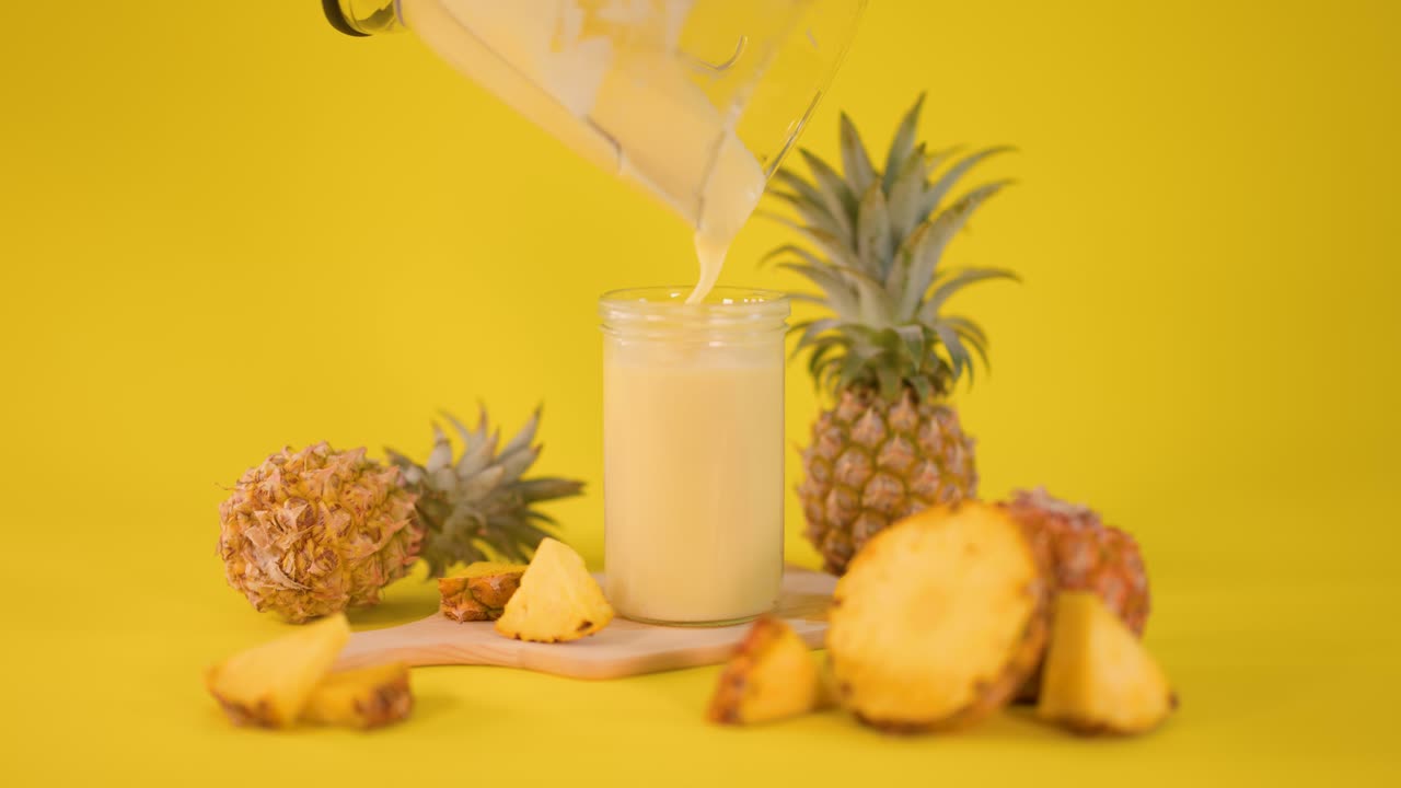 Creamy pineapple smoothie poured into glass, surrounded by fresh fruit, bright yellow background, static shot