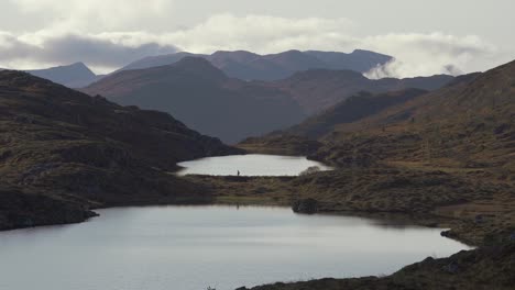 Person-crossing-a-lake-in-the-distance,-set-in-a-beautiful-mountain-and-lake-scenery