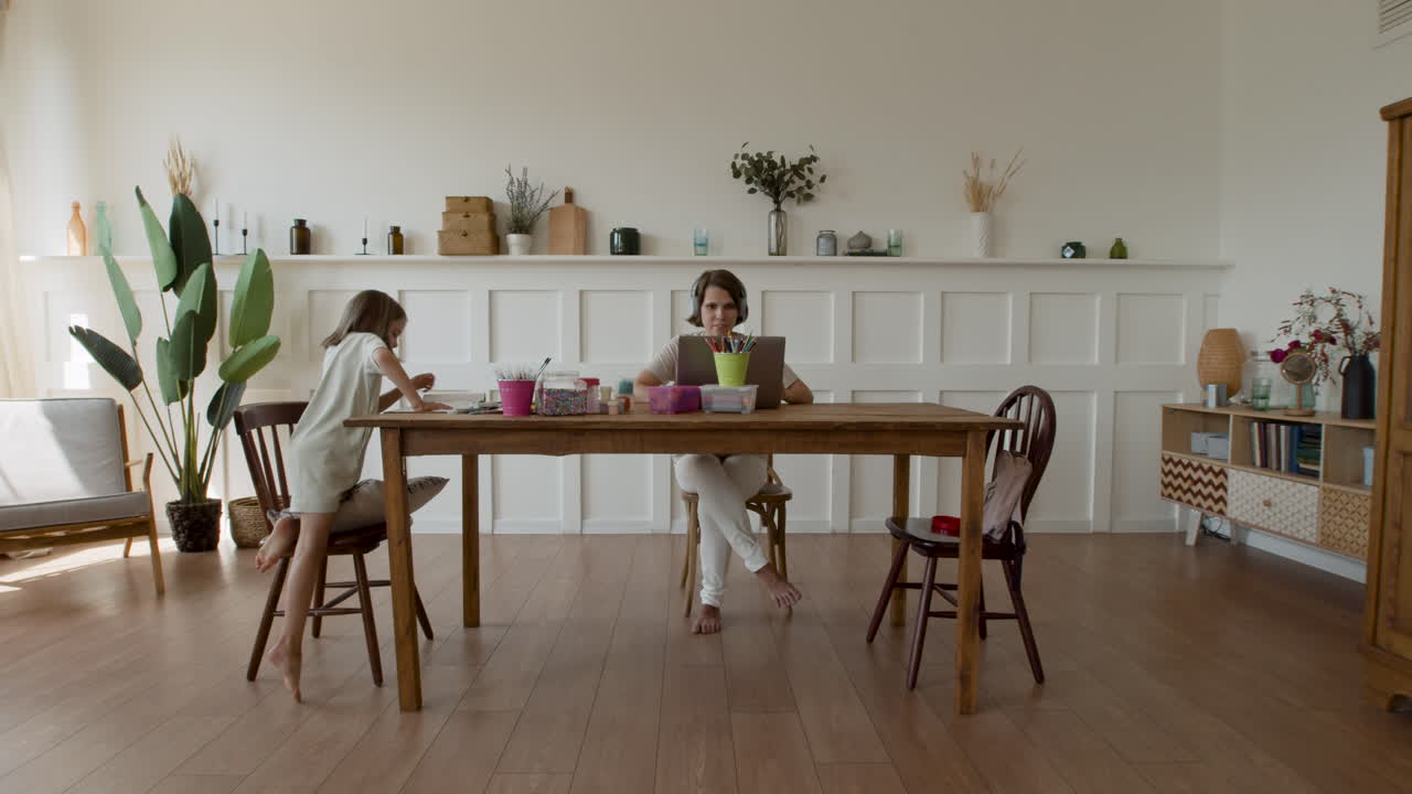 plano general de una madre que trabaja en casa haciendo una videollamada cuando su hija entra y se sienta a su lado para hacer su tarea
