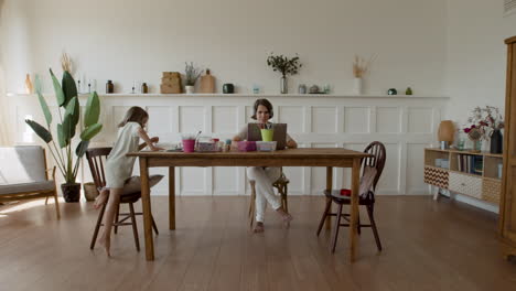 wide shot of a mother working at home making a videocall when her daughter comes in and sits down next to her to do her homework