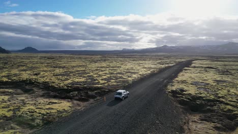 una toma panorámica de un suv en un camino de grava en islandia, rodeado de montañas y terreno cubierto de musgo, filmado desde una vista aérea, mostrando el hermoso paisaje