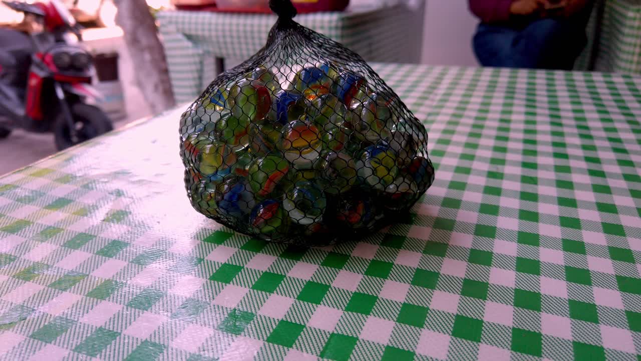 Camera slowly pulls back from a mesh bag filled with colorful glass marbles placed on a green checkered table
