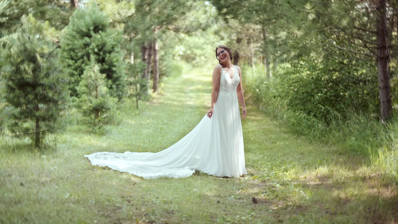 Static full-body view as short bride with glasses smiles, admiring her dress train