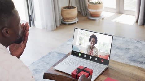 Hombre-Afroamericano-Sosteniendo-Un-Regalo-Rojo-Y-Usando-Una-Computadora-Portátil-Con-Una-Mujer-Afroamericana-En-La-Pantalla