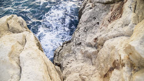 Looking-down-at-blue-ocean-sea-water-waves-crashing-on-steep-rocky-tan-cliffs,-Malta,-zoom-out