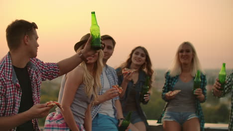 a boy raises a toast to a friend's birthday in friends company on the roof. they clink beer and eat pizza.