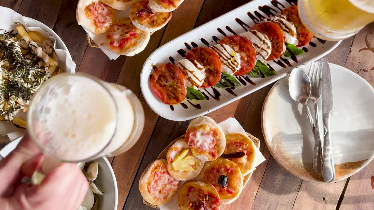 A vibrant beachside meal with bruschetta, pizza, and clams, highlighted by a toast with frothy beers under warm lighting