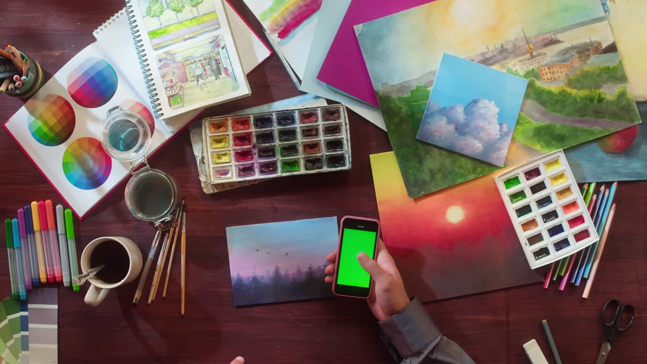 Artista pintando un paisaje con acuarelas y usando un smartphone con pantalla verde