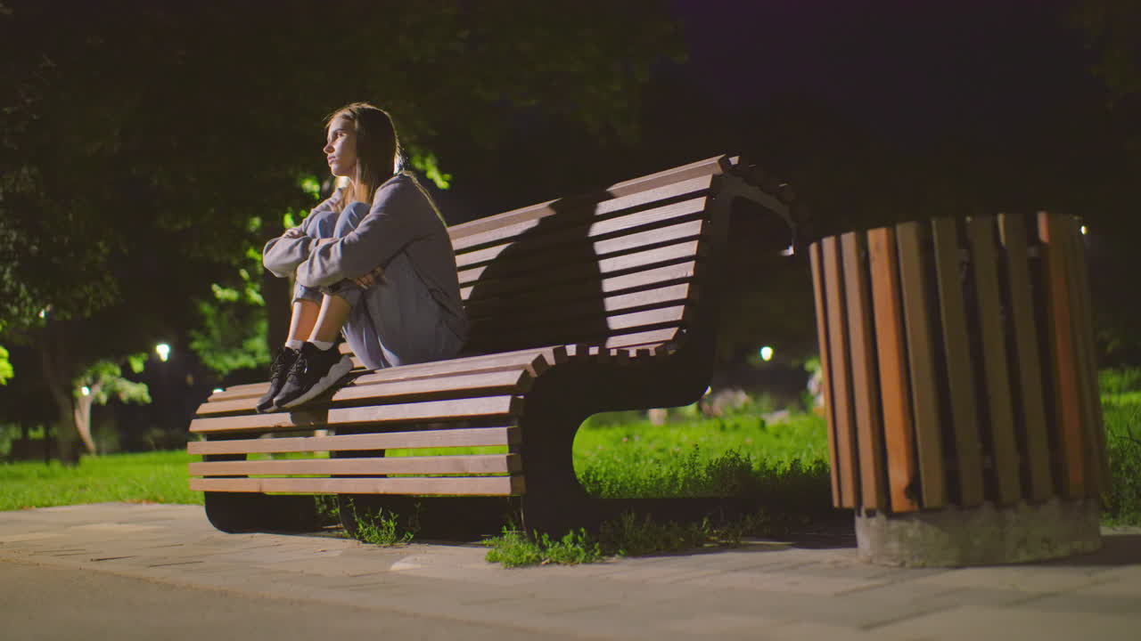 Lady seated on park bench at night with arms crossed over knees, gazing thoughtfully around, illuminated by soft park lighting, surrounded by greenery and shadows