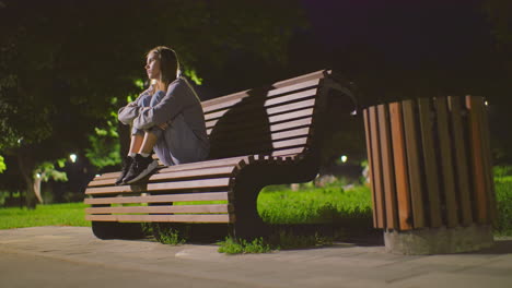 lady seated on park bench at night with arms crossed over knees, gazing thoughtfully around, illuminated by soft park lighting, surrounded by greenery and shadows