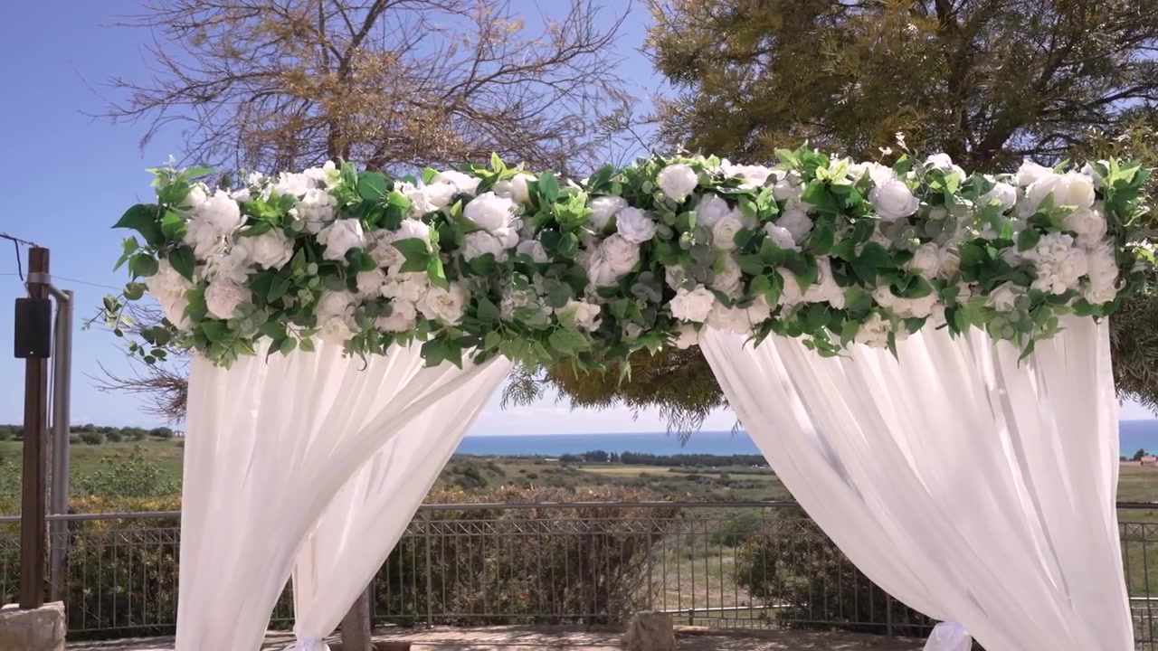Un arco floreale fatto di fiori bianchi e foglie verdi si trova in una location per matrimoni all'aperto. L'oceano è visibile sullo sfondo sotto un cielo azzurro e limpido durante il giorno