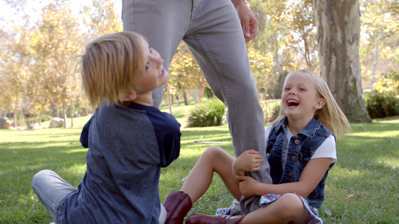 Two young kids playing on their dad's legs in a park
