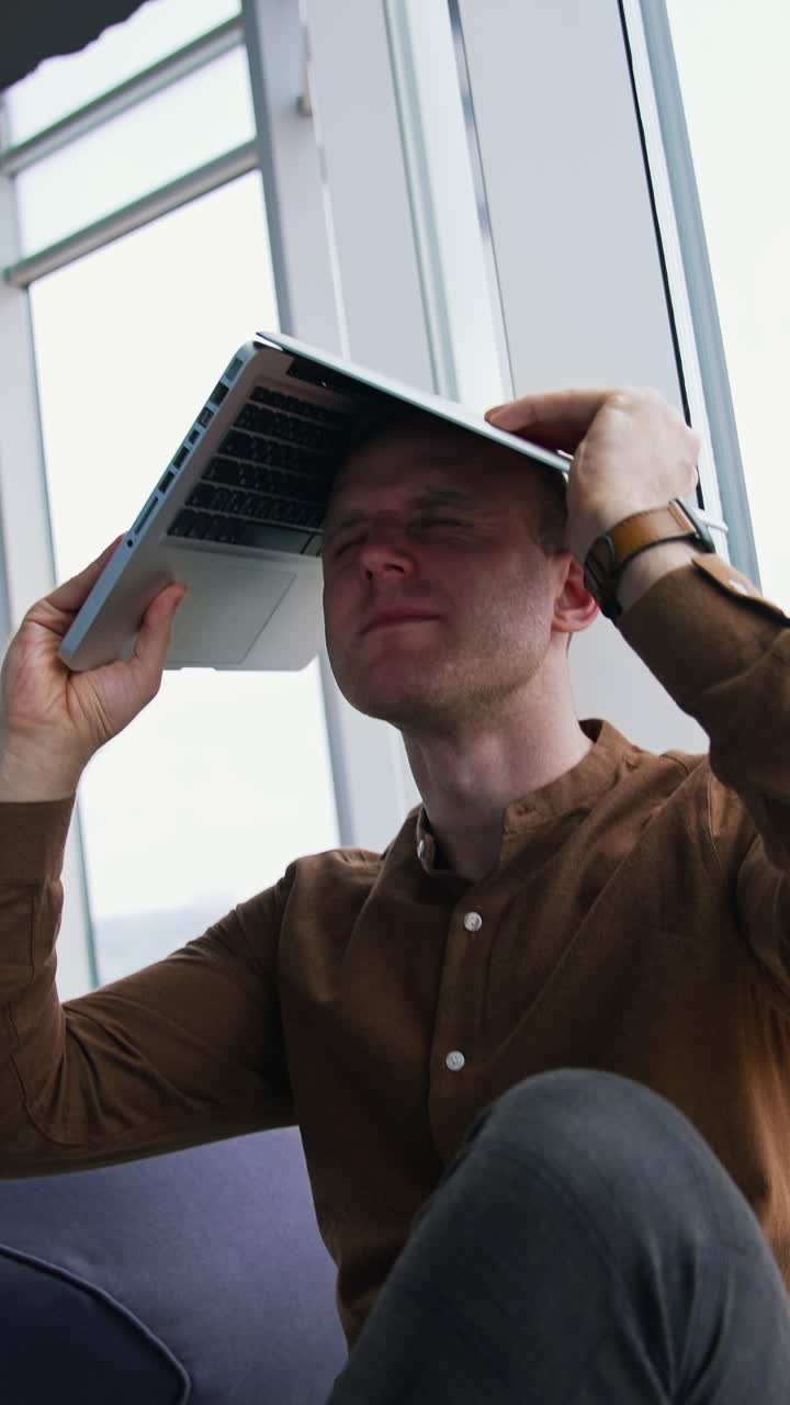 Tired businessman with a laptop on his head. Man is sitting in the office by the large window and holding a laptop over the head. Problems in business. Vertical video