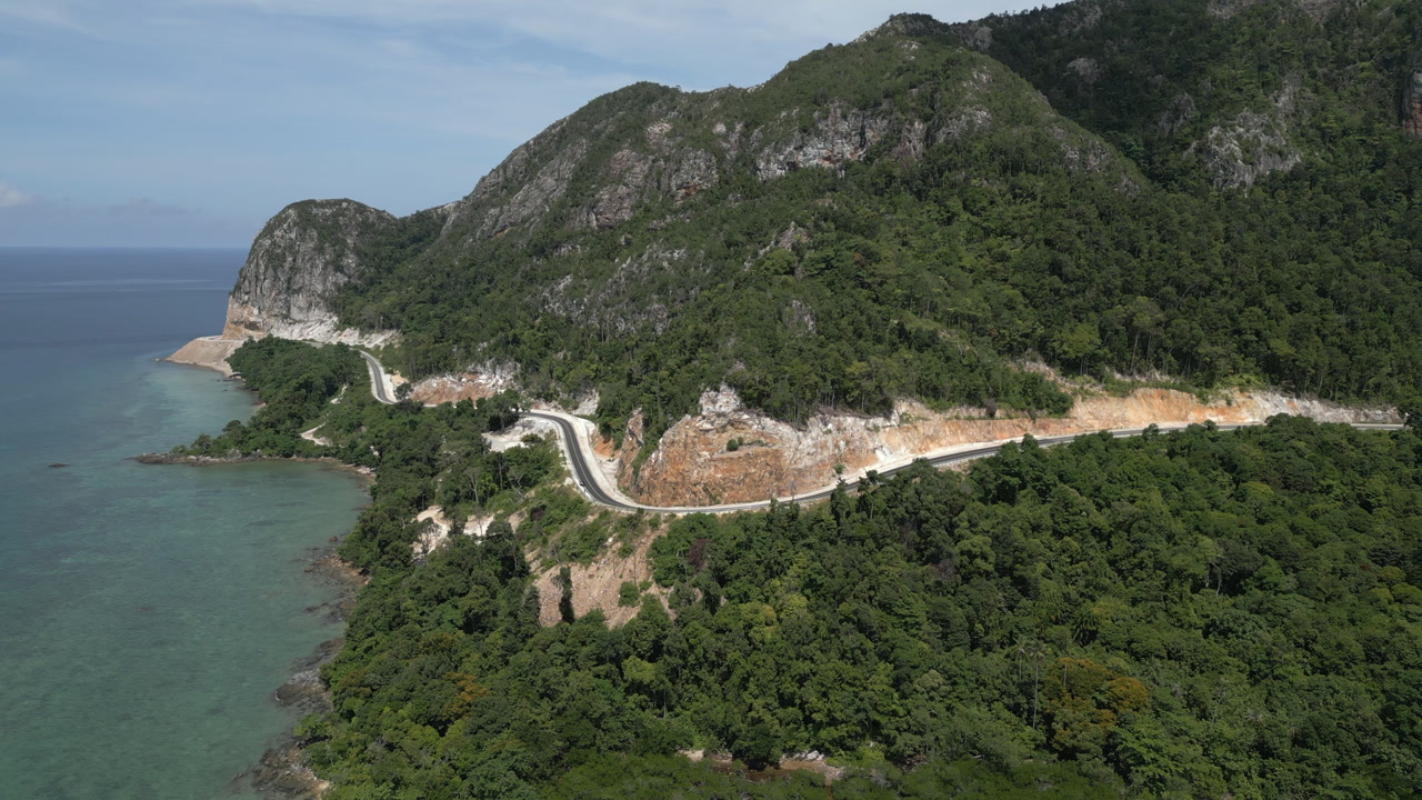 Aerial drone footage of a winding mountain road situated between steep rocky cliffs and the ocean. Captured at Bukit Tekol, featuring the dramatic coastline of South Natuna, Indonesia.