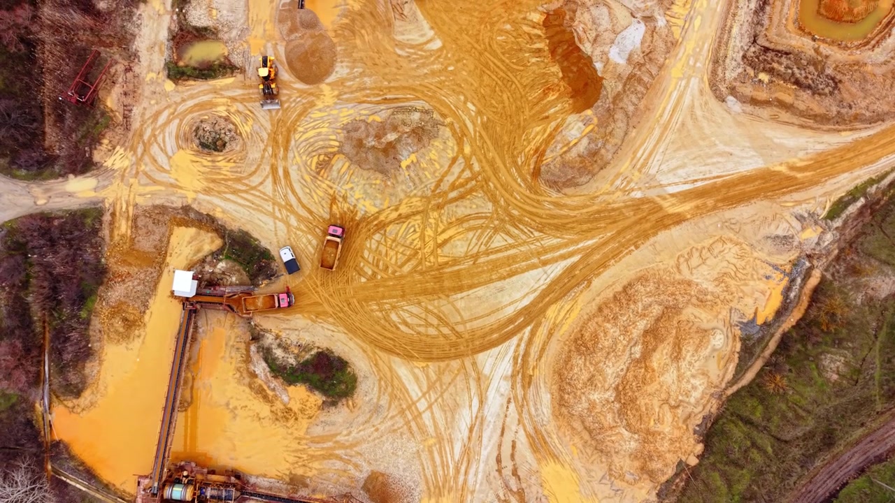 Overhead view of sand quarry with mining trucks. Top down perspective reveals orange sand piles, tire tracks, and a conveyor structure.