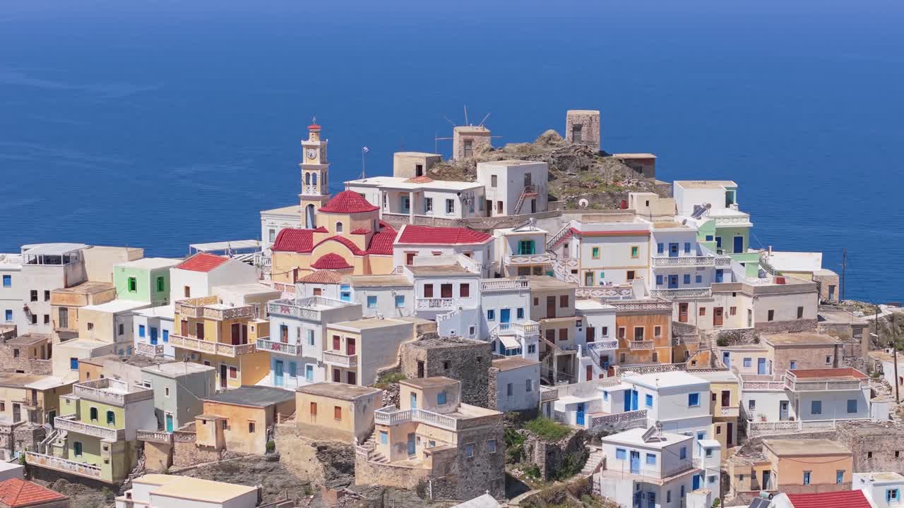 Drone orbit around a picturesque village in Karpathos, Greece, showcasing the main square, church, blue sky, and colorful traditional houses of this beautiful Aegean island