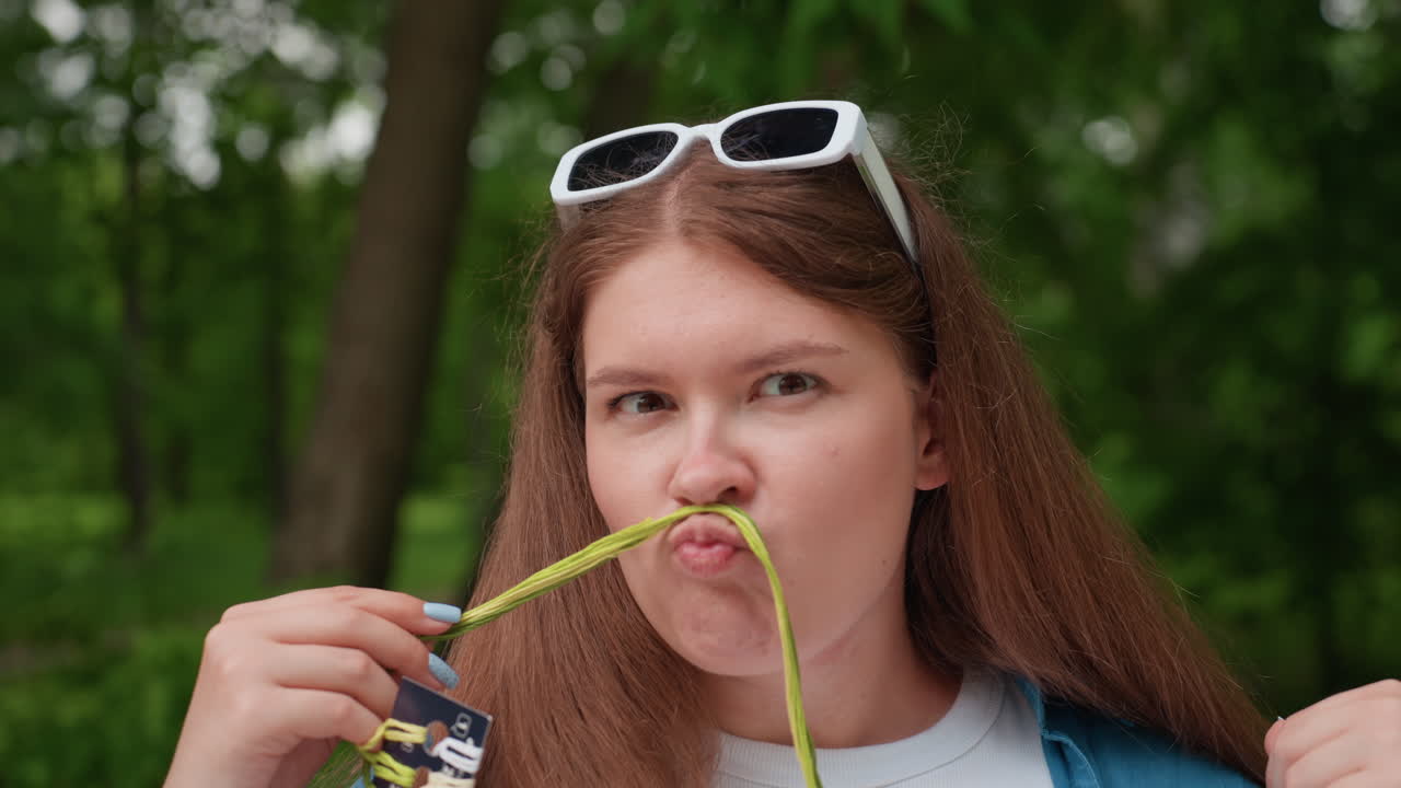 Close up of funny lady sitting on bench outdoors holding green thread under nose making mustache face while turning eyes humorously, surrounded by lush green park scenery