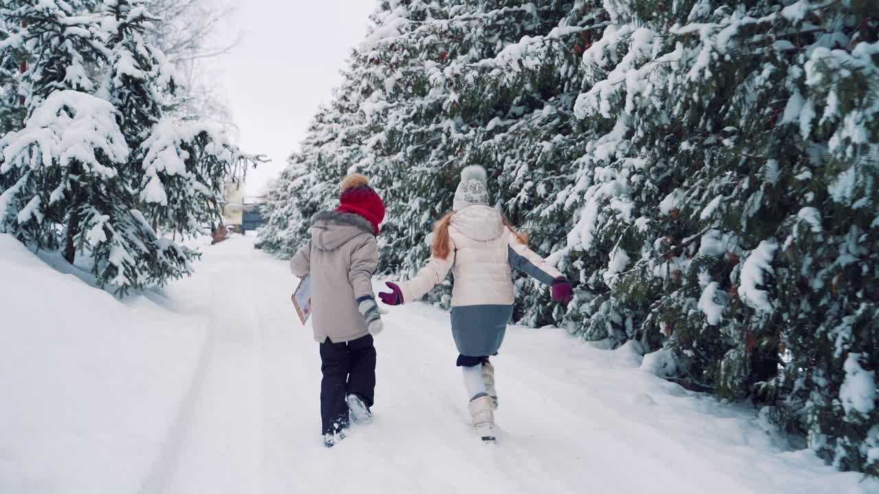 Children with their backs are running along the snowy road and holding each other's hands on the background of coniferous forest on two sides. The little boy is holding a brown envelope in his hand