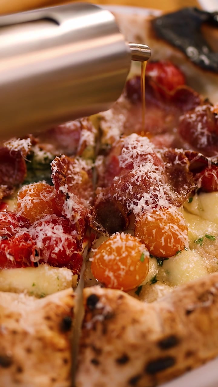 Primo piano di olio d'oliva che cola su pizza fresca con prosciutto e pomodorini.