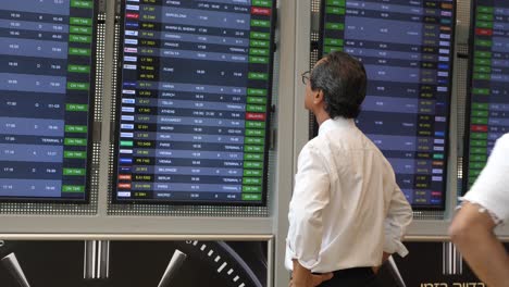 Un-Hombre-De-Negocios-Japonés-Se-Encuentra-En-El-Aeropuerto-Ben-Gurion-En-Israel,-Mirando-El-Tablero-De-Llegadas-Y-Salidas.