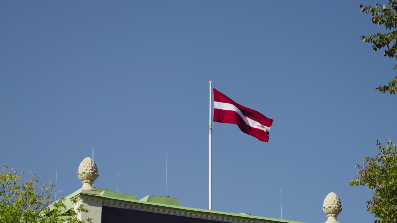 Handheld shot of Latvian flag waving on top of building against blue sky