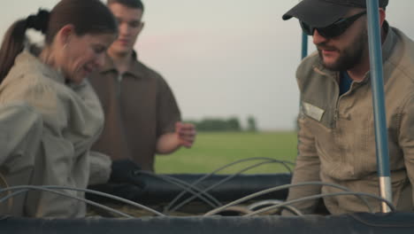 close up of couple in gloves lowering metal burner assembly from hot air balloon basket as young boy watches intently against soft dusk sky highlighting detailed manual setup teardown process