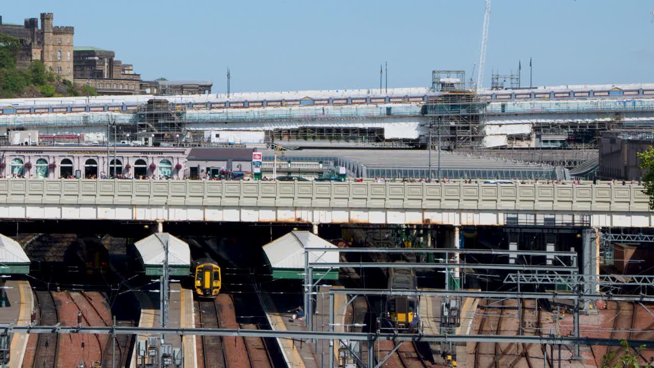 Yellow-fronted train enters busy city station under clear daylight, historic buildings in background