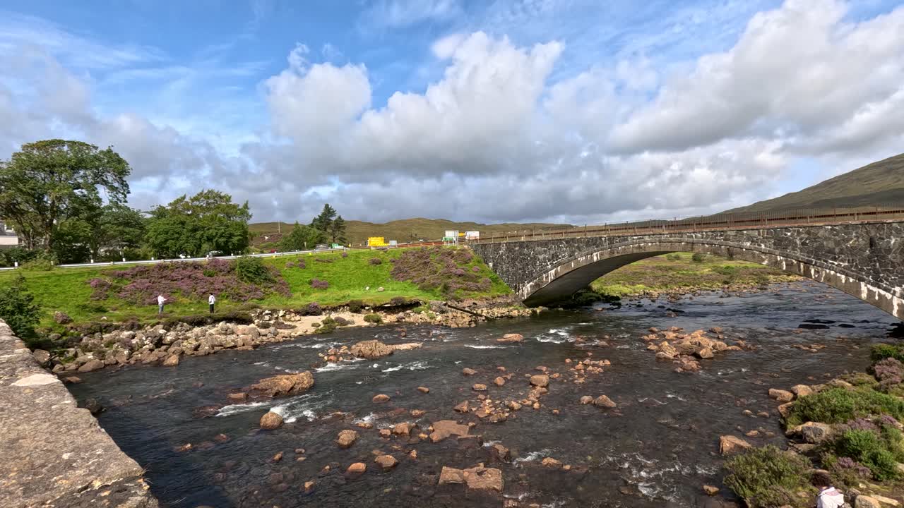 Wide-angle view of several people walking along a rocky stream beneath an old stone bridge, with lush greenery and hills under bright daylight