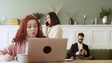 a redhead woman working on laptop computer while behind her a man using mobile phone sitting on the sofa and an girl talking on phone walking in the room