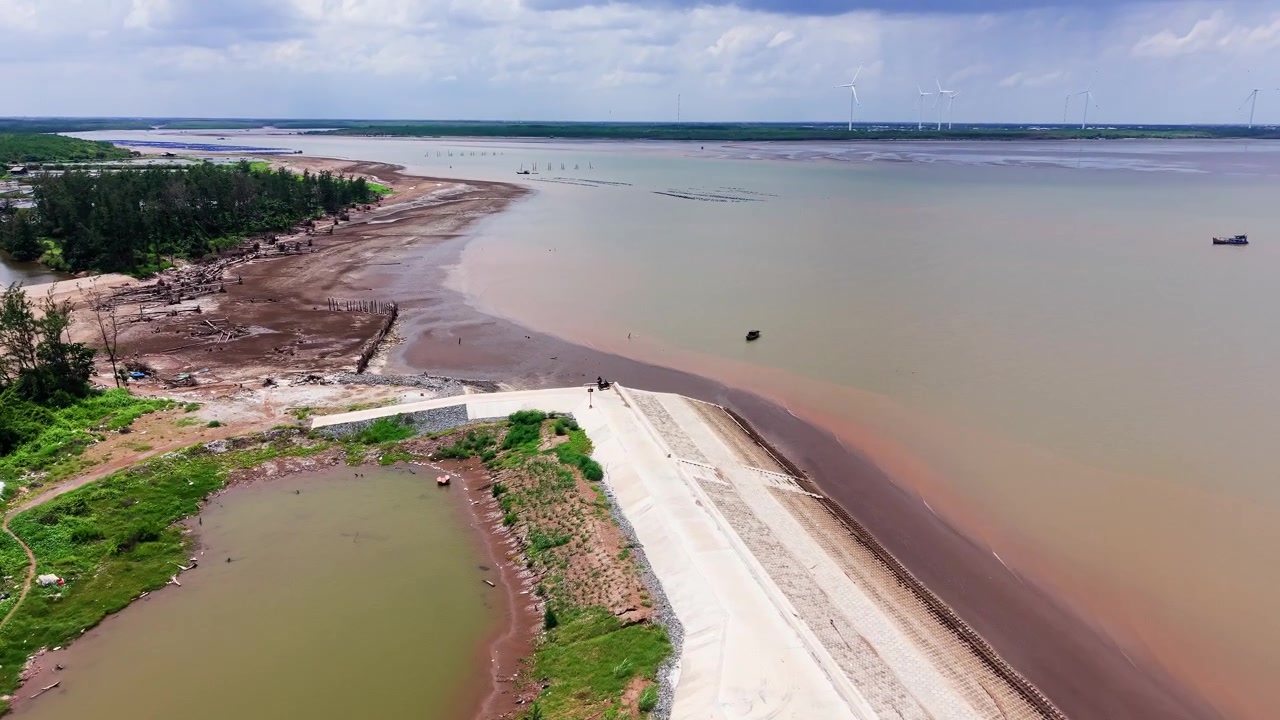 Visitatori camminano lungo la riva della spiaggia di Con Nhan a Vinh Long, Vietnam. Sono visibili barche nell'acqua. Il sole splende sopra mentre passano le nuvole. Vegetazione verde costeggia l'area della spiaggia