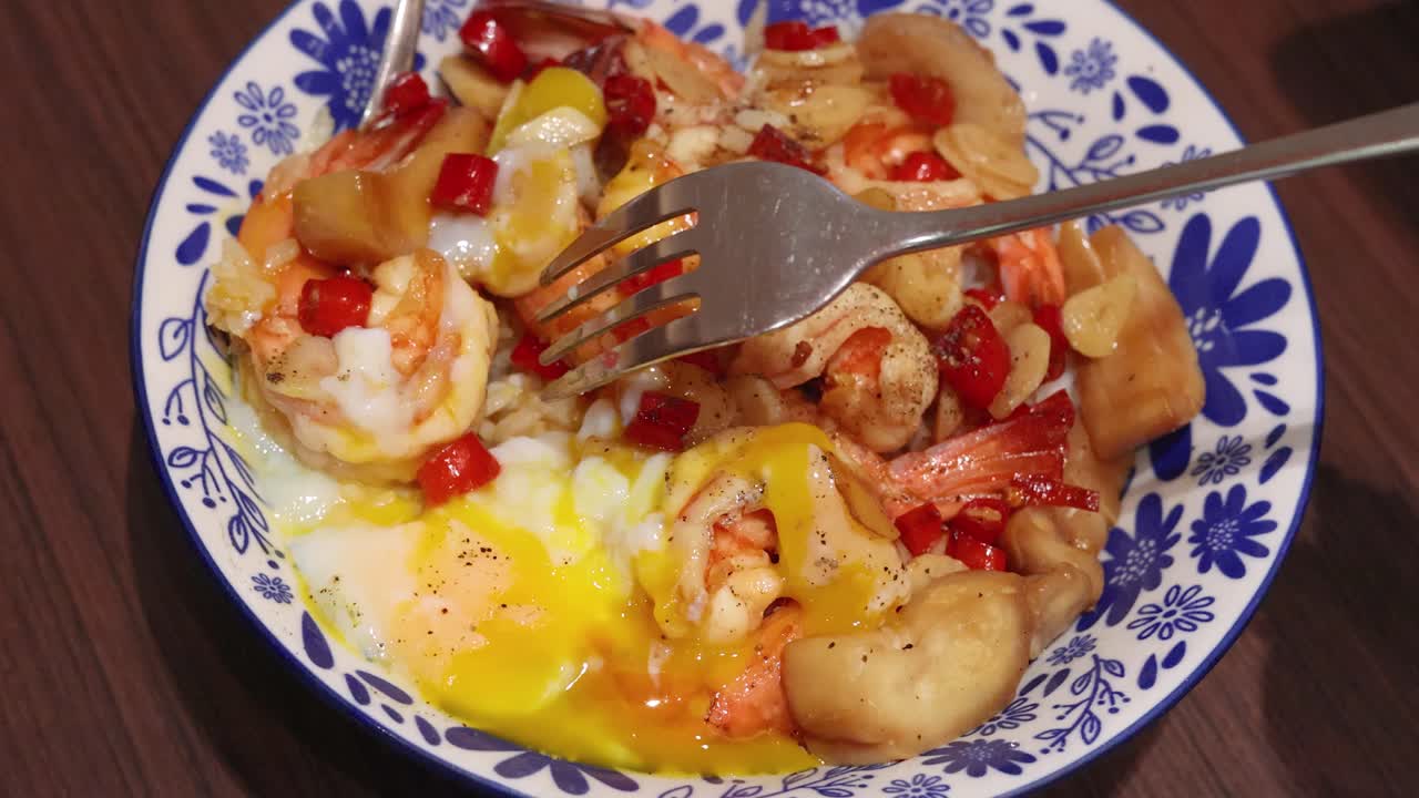A fork stirs shrimp, garlic, chili, and rice with runny egg yolk in a patterned bowl under warm indoor lighting, highlighting Thai cuisine textures