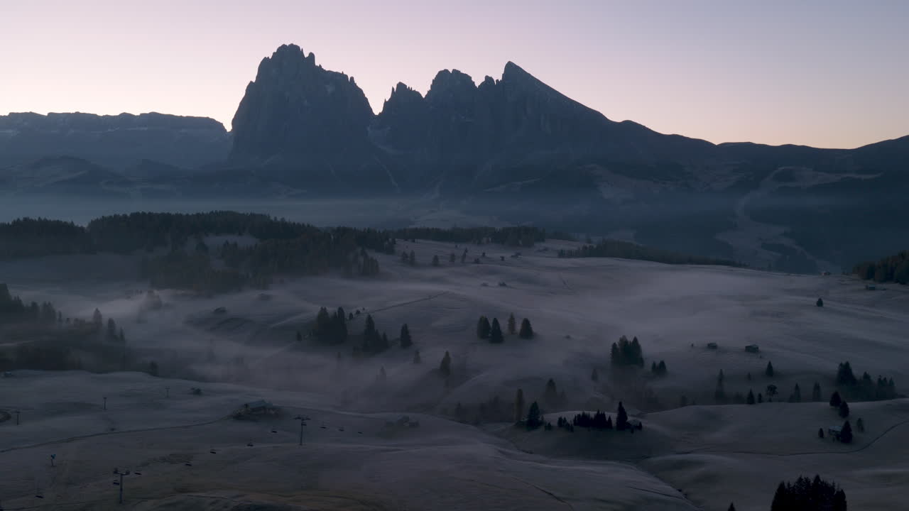 Filmisk luftfoto af frostklædte Alpe di Siusi-enge med spredte bjerghytter og morgentåge, dramatiske Dolomitter-toppe i de italienske alper ved solopgang, fredfyldt natursceneri