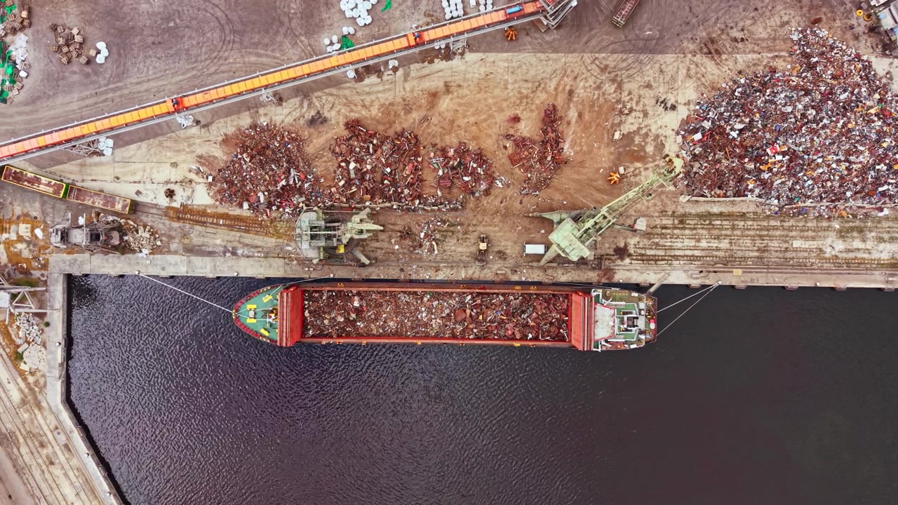 Grande nave da carico attracca in un porto industriale per scaricare rottami metallici durante le ore pomeridiane, circondata da cumuli di metallo e macchinari pesanti