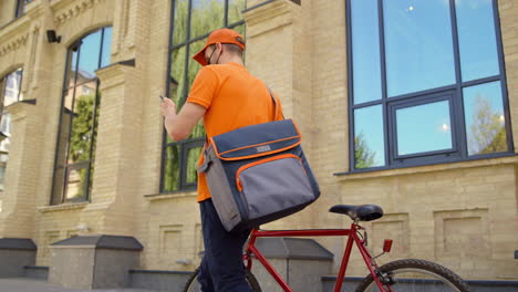 masked courier using mobile phone outdoors. uniform man leading bicycle street.