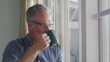 Happy-caucasian-man-wearing-glasses,-looking-through-window-and-drinking-coffee