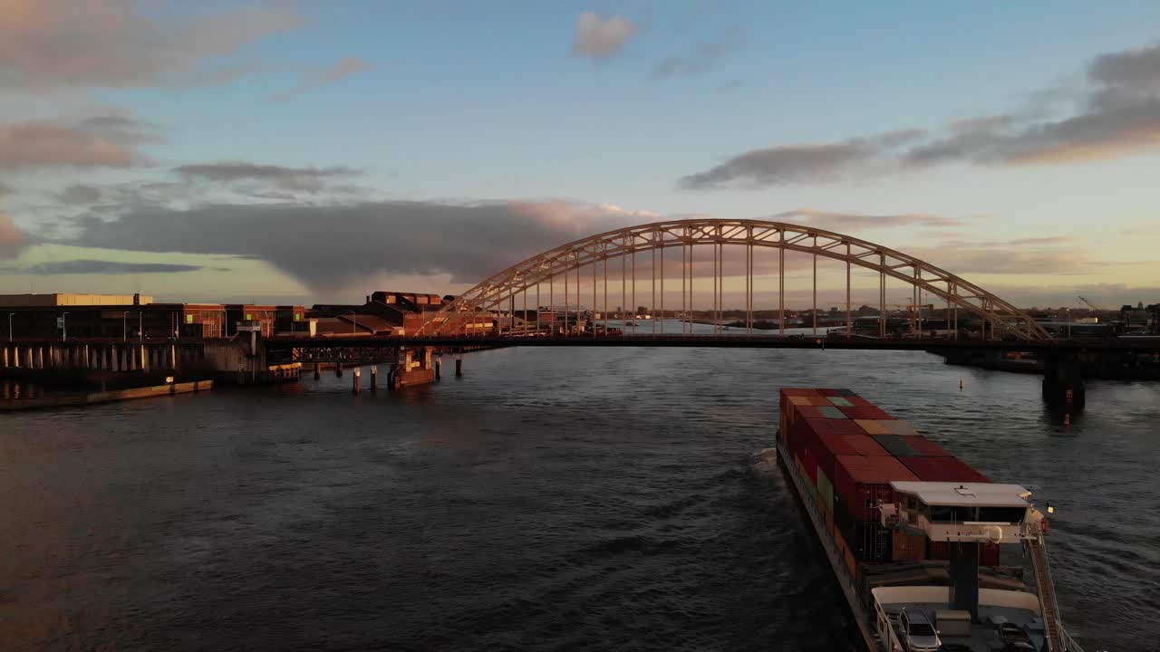 Fully loaded container barge Navigating towards a bridge over the dutch river Noord  on a partly cloudy day during golden hour. Drone dolly shot