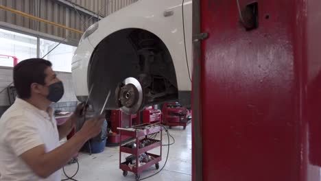 dirty latin male mechanic removing the fender liner of a white car without wheel with two screw drivers at a workshop station in mexico latin-america